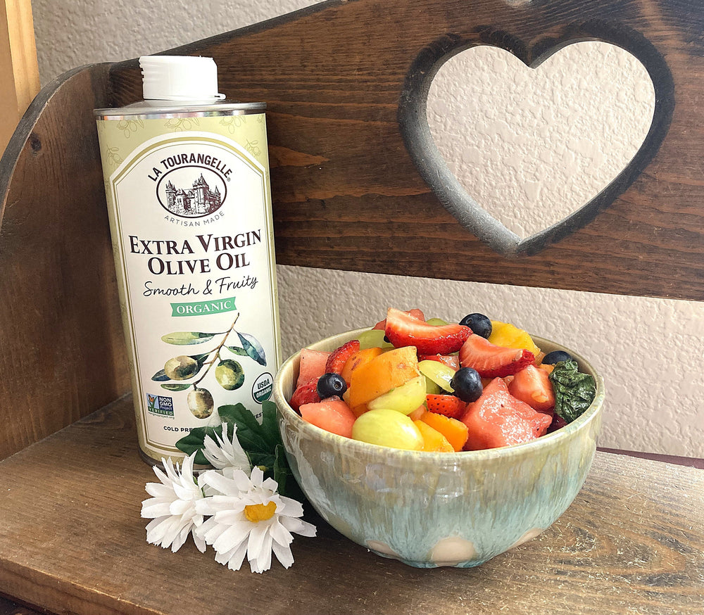 A fruit salad in a small bowl, sitting on a wooden shelf with La Tourangelle's extra virgin olive oil can in the background.